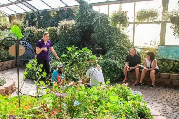 A staff member in a purple shirt talks with visitors seated on the low garden wall, surrounded by flower beds under the glass roof.