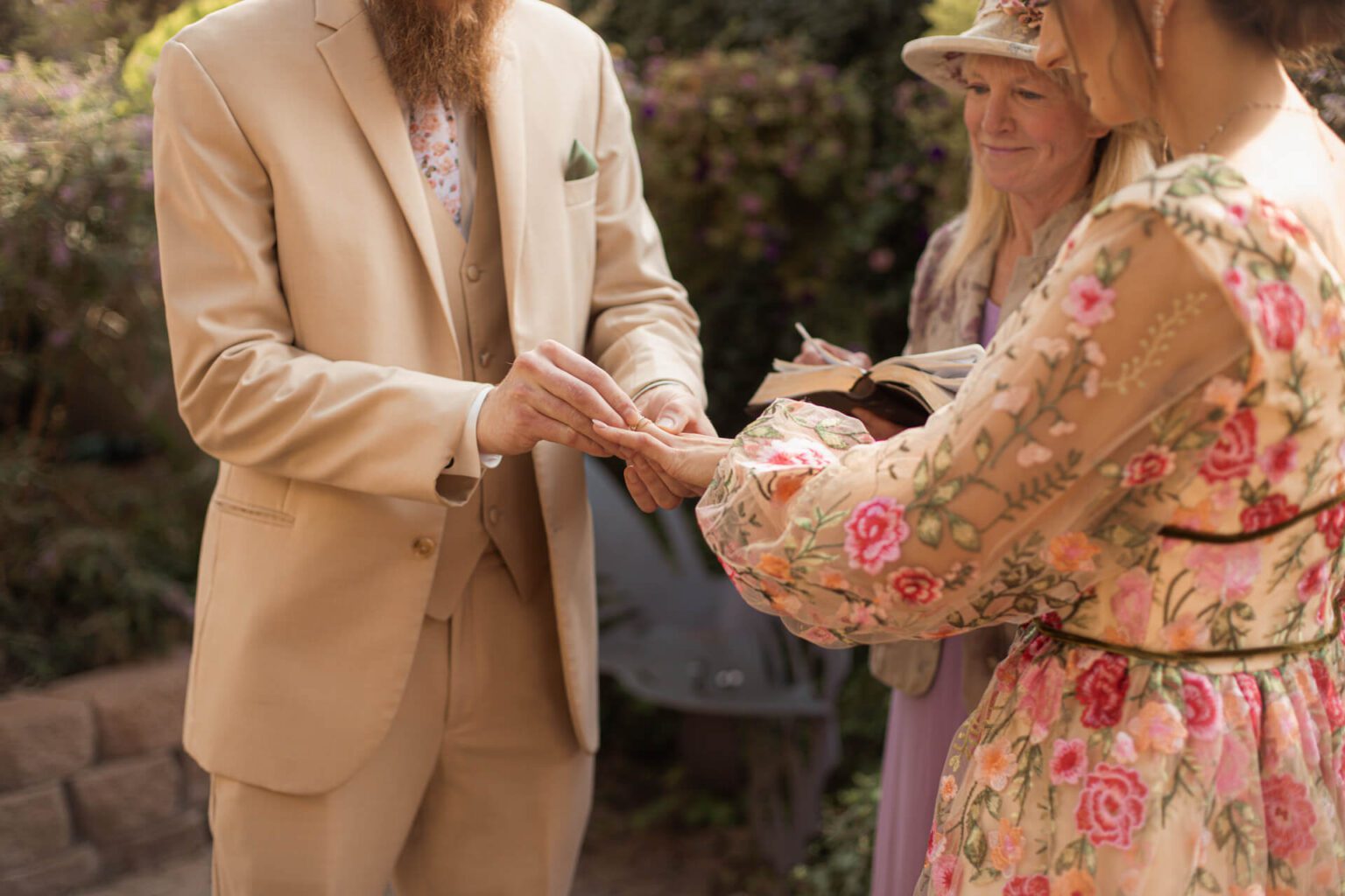 A groom in a tan suit slides a ring onto the bride’s finger while the officiant reads, in front of flowering shrubs.